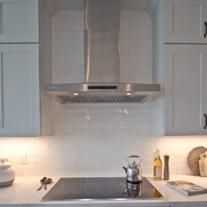 A chrome range hood is center of image with white shaker style wall cabinets on each side. A white, glazed ceramic tile backsplash is set behind