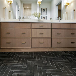 modern transitional elegance Primary bathroom floor featuring Ragno Rewind Brick 3x11 Peltro tile installed in a herringbone pattern beneath a light natural wood vanity topped with white quartz, expertly designed by Coyle Carpet One in Madison, WI.