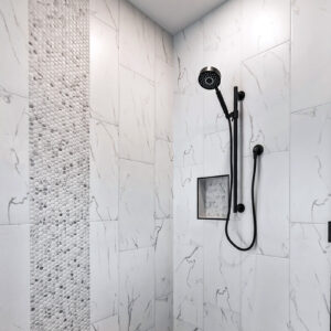 The primary bathroom shower features Emser Contessa 12x24 Oro matte tile on the walls, complemented by Daltile Famed Penny Round Mosaic in Iconic for the floor, creating a luxurious and modern spa-like retreat.